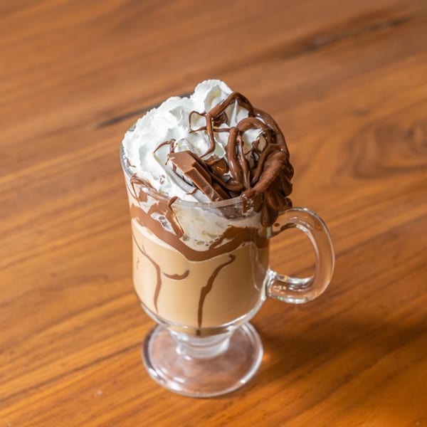 A glass mug of coffee with whipped cream, chocolate drizzle, and chocolate bar pieces on a wooden table.