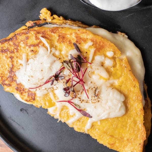 Close-up of a golden Venezuelan cachapa folded over melted white cheese, garnished with red microgreens.