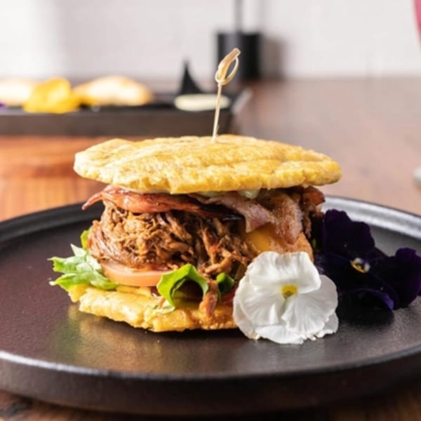 A Venezuelan patacon with shredded beef, lettuce, tomato, and cheese, garnished with edible flowers.