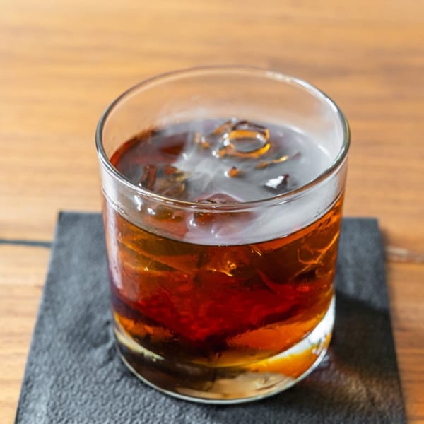 A glass of dark iced tea or cocktail on a black coaster, with a wooden table background.