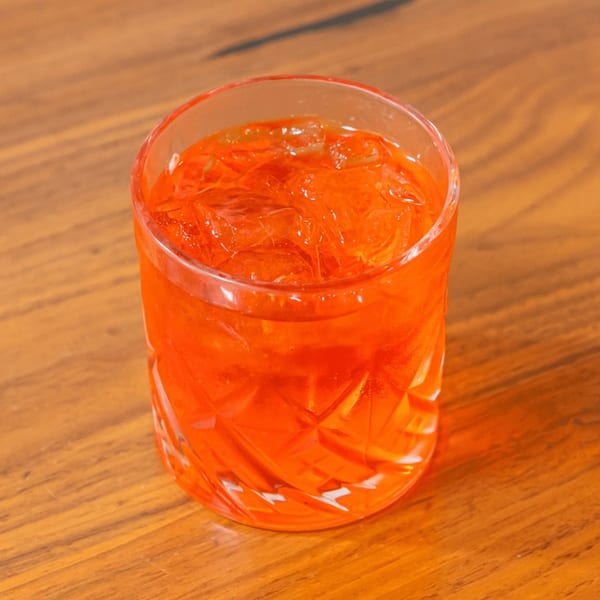 A vibrant orange cocktail with ice in a textured glass sits on a warm wooden table.