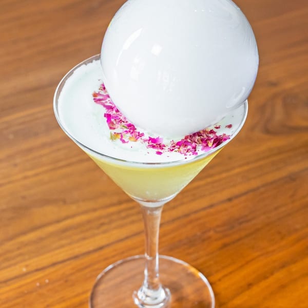 A martini glass with a yellow drink, white foam, pink petals, and a large white bubble.
