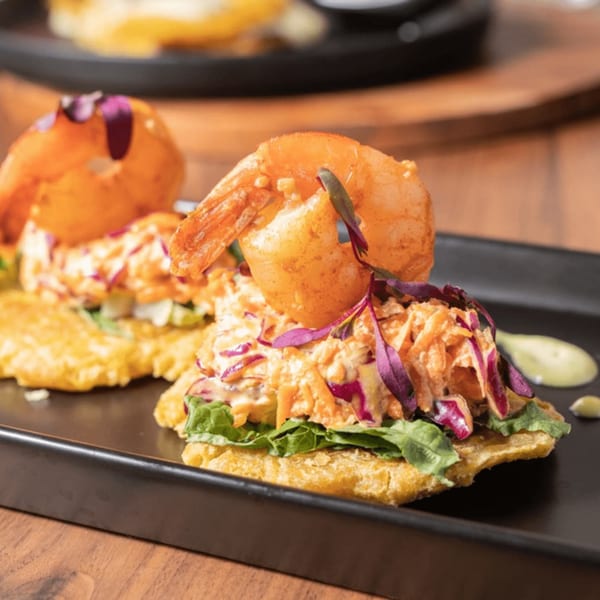 Two Venezuelan patacones topped with shrimp, coleslaw, and purple garnish on a black plate.