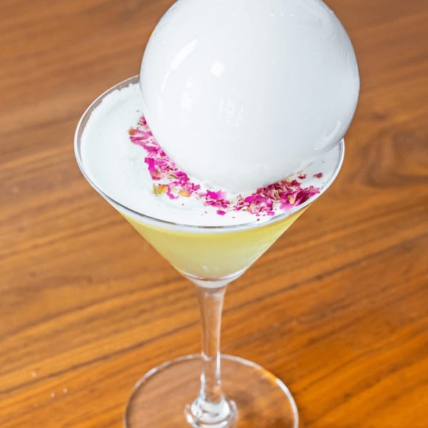A martini glass with a yellow drink, white foam, pink petals, and a large white bubble.