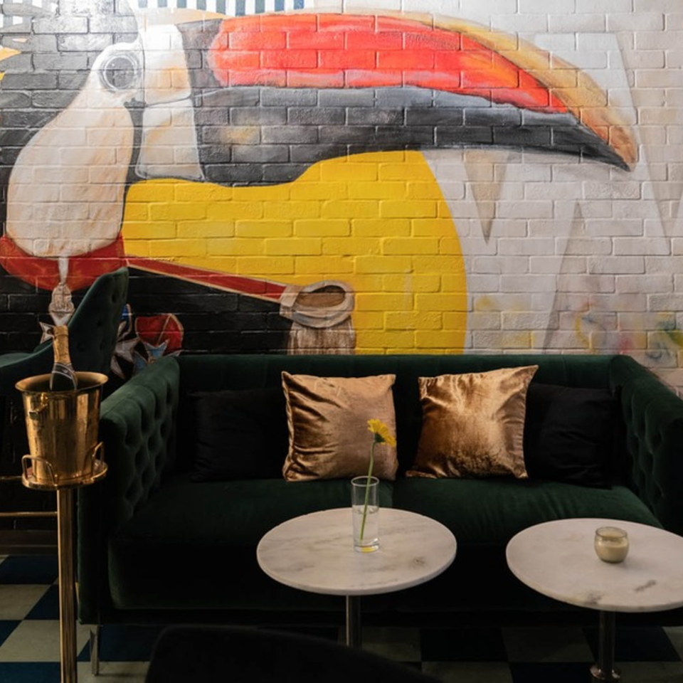A vibrant toucan mural on a brick wall above a dark green sofa with gold pillows and marble tables.