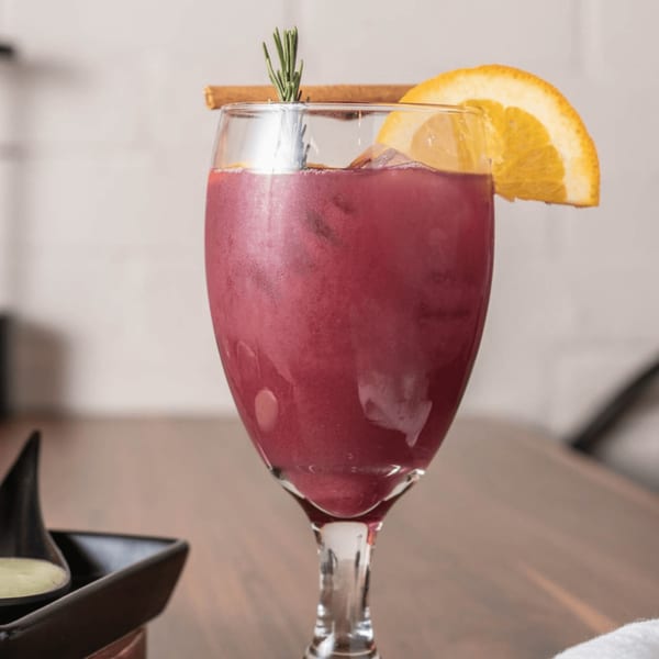 A vibrant red drink in a wine glass, garnished with an orange slice, cinnamon stick, and rosemary.