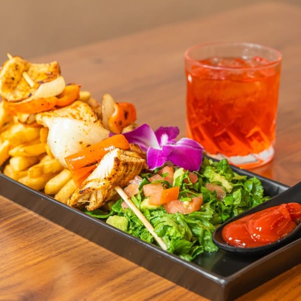 Venezuelan chicken skewers with fries, salad, and a red drink on a black tray.