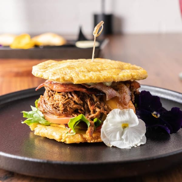 A Venezuelan patacon with shredded beef, lettuce, tomato, and cheese, garnished with edible flowers.