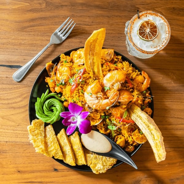 Venezuelan shrimp and rice dish with plantains, avocado, and a purple orchid, served with a cocktail.
