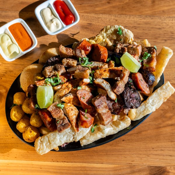 A large platter of mixed grilled meats, sausages, potatoes, and fried plantains, with dipping sauces.